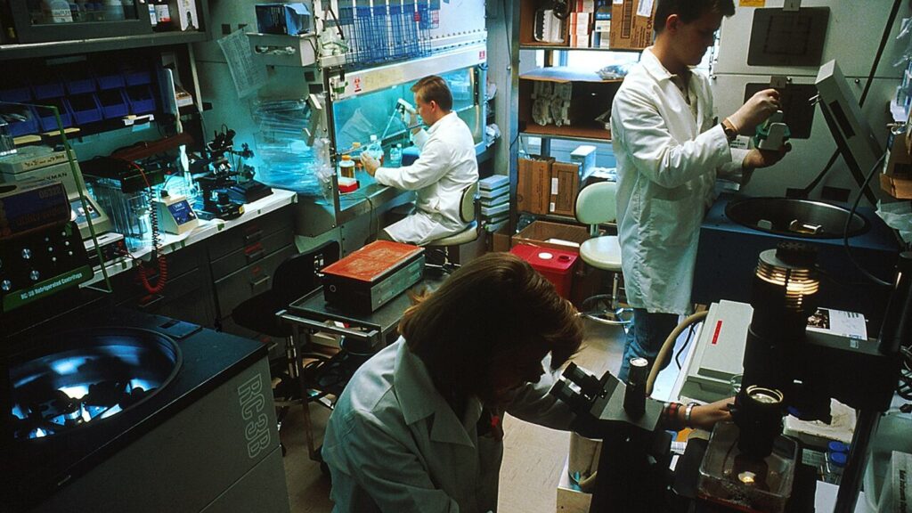Three scientists in lab coats working at benches and a microscope in a research laboratory
