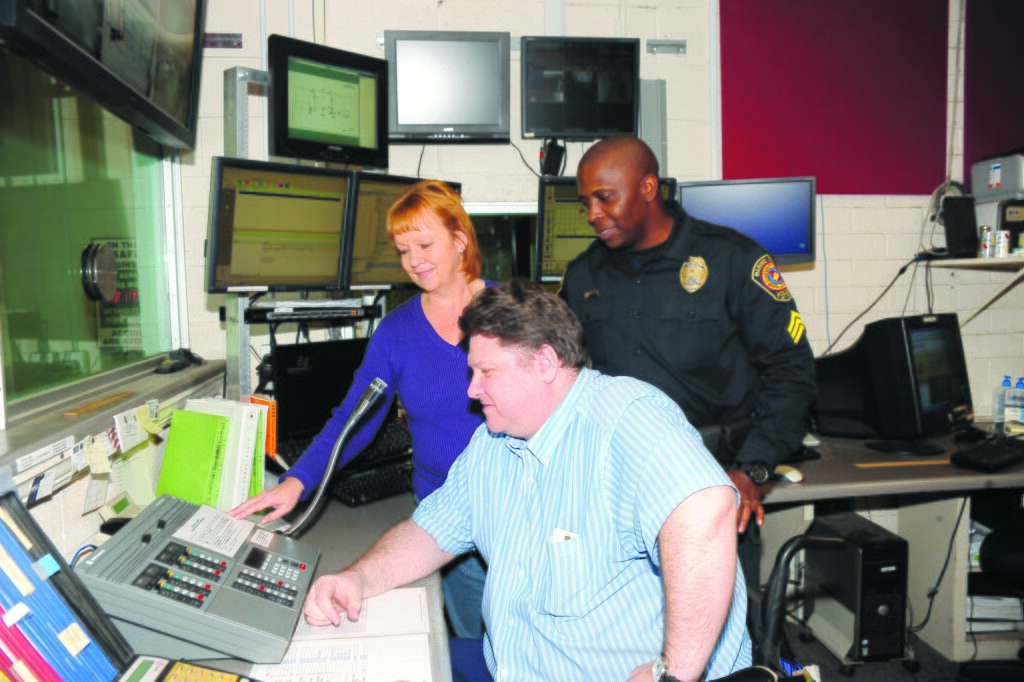 Emergency services dispatcher seated at a police radio dispatch console while a radio manager and a patrol sergeant look on